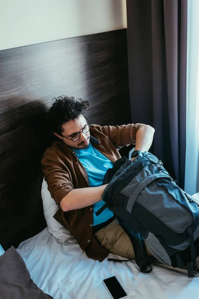 男性の観光客がホテルでのスマート フォンが付いているベッドに座ってバックパックから何かを取る — ストック写真