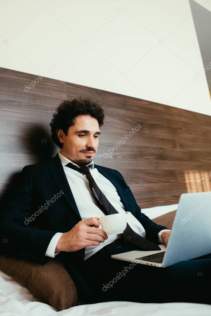 adult businessman in suit holding cup of coffee and teleworking on laptop in hotel room