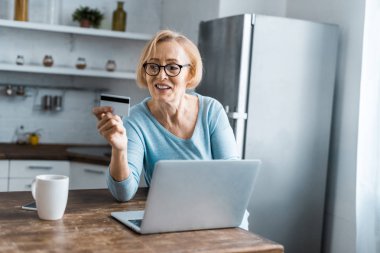 smiling senior woman holding credit card and using laptop while doing online shopping at home clipart