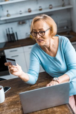 kredi kartı tutan ve evde online alışveriş yaparken dizüstü kullanarak üst düzey kadın seçici odak