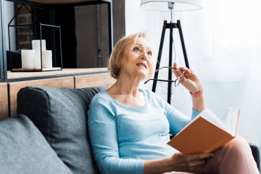 kanepede oturan, gözlük tutan ve uzak evde kitap okurken seyir dalgın kadın kıdemli 