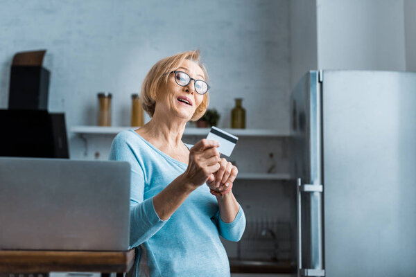 surprised senior woman in glasses holding credit card near laptop at home with copy space