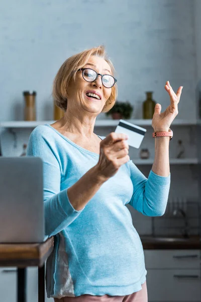 Zaskoczony Starszy Kobietę Okularach Gestem Ręką Posiadania Karty Kredytowej Pobliżu — Zdjęcie stockowe