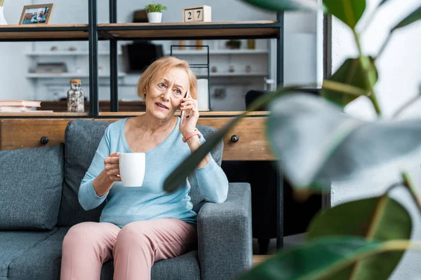 Mulher Sênior Confuso Com Xícara Café Sentado Sofá Falando Smartphone — Fotografia de Stock