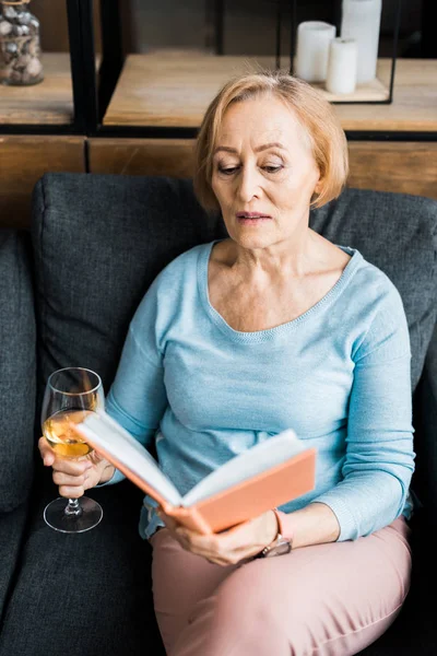 Mulher Sênior Sentado Sofá Segurando Copo Vinho Livro Leitura Casa — Fotografia de Stock