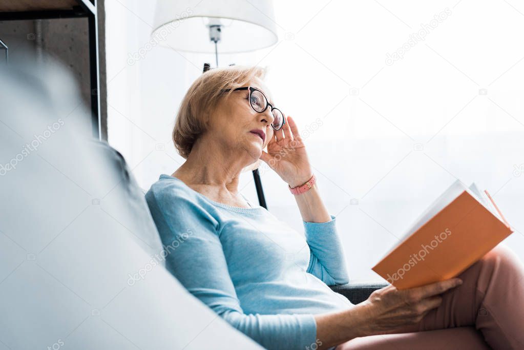 selective focus of senior woman in glasses sitting and reading book at home 