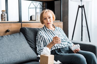 sick senior woman looking at camera and holding bottle of medication with glass of water 