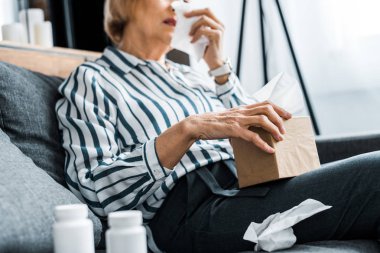 partial view of sick senior woman with runny nose holding tissue box at home