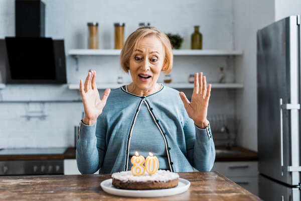 excited senior woman sitting and looking at cake with '80' sign on top during birthday celebration