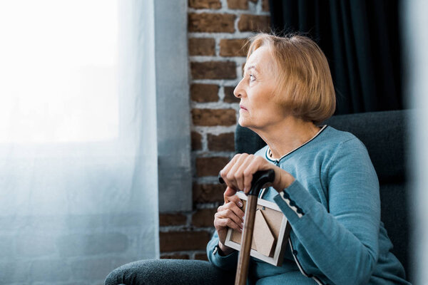 upset senior woman sitting with walking stick and holding picture frame at home