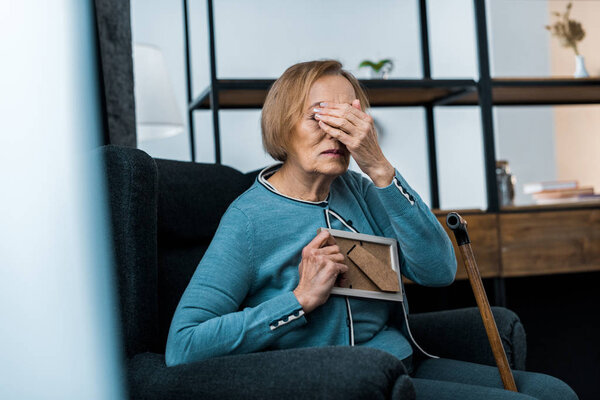 grieving senior woman sitting in armchair, covering face with hand and crying while holding picture frame