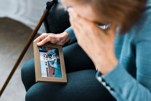 пожилая женщина закрывает лицо рукой и плачет, глядя на рамку с мужчиной на фотографии
