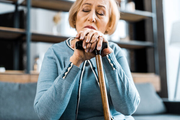 sad senior woman with eyes closed leaning on walking stick at home
