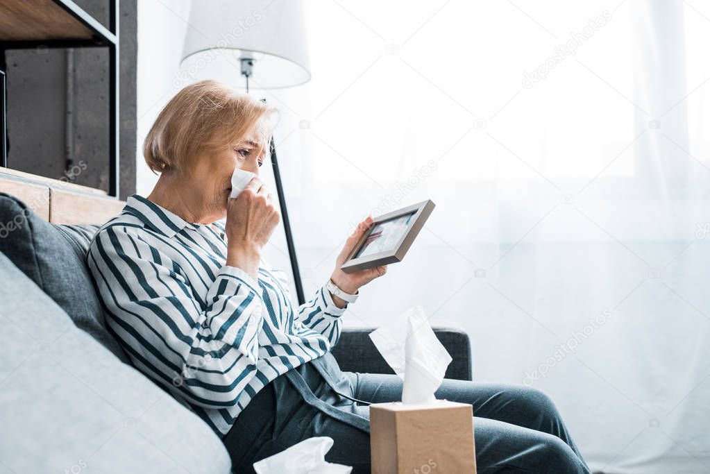 depressed senior woman crying and wiping face from tears with tissue while looking at picture frame 
