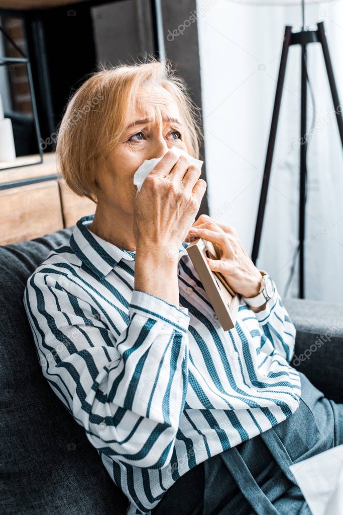 sad senior woman wiping face from tears with tissue and crying while holding picture frame