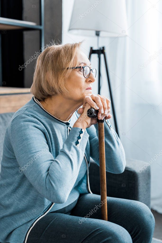 depressed senior woman in glasses sitting with walking stick and looking away at home