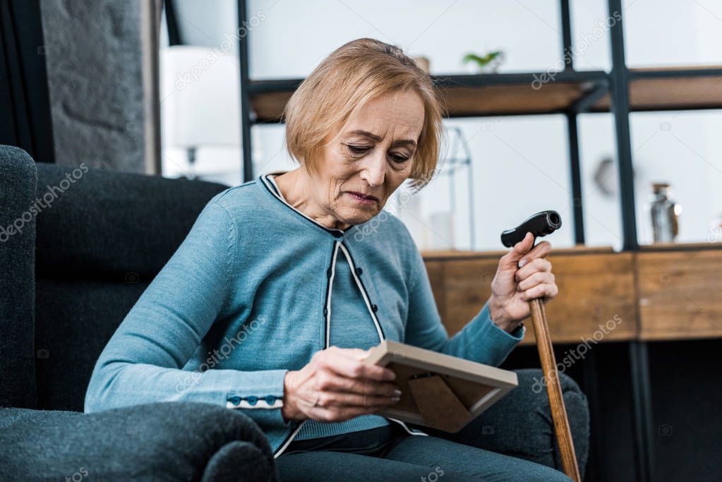 sad senior woman sitting with walking stick and looking at picture frame