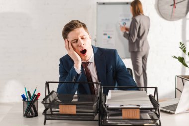 selective focus of tired man yawning near document trays with lettering with female coworker on background, procrastination concept clipart
