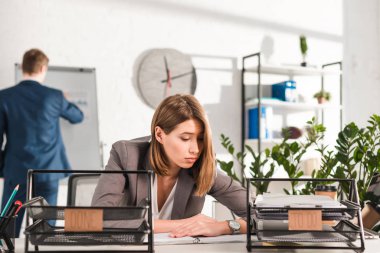 selective focus of sleepy businesswoman sitting at desk near document trays with lettering, procrastination concept clipart