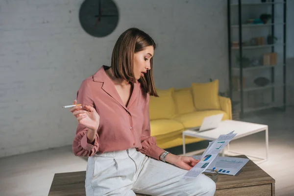 Attraktiv Kvinna Håller Cigarett Och Tittar Tabeller Och Diagram Hemma — Stockfoto