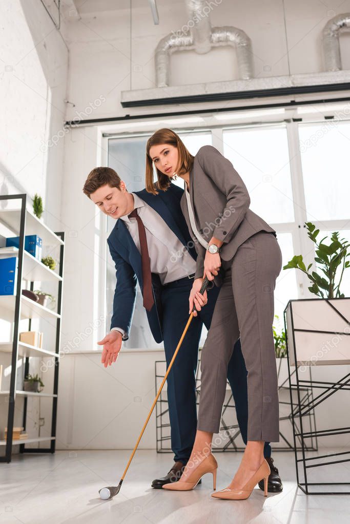 handsome businessman gesturing while looking at ball near female coworker playing mini golf in office 