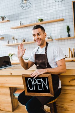 handsome cashier in apron sitting on chair near wooden bar counter, holding chalkboard with open lettering and waving with hand in coffee house clipart