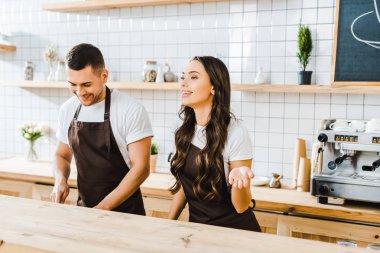 attractive cashier standing behind wooden bar counter and smiling wile barista working in coffee house clipart