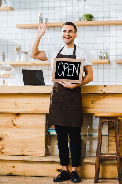 handsome cashier standing near wooden bar counter, holding chalkboard with open lettering and greeting in coffee house