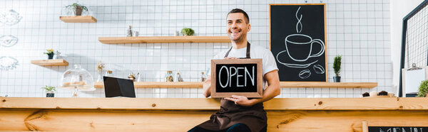 handsome cashier in brown apron sitting on chair near wooden bar counter and holding chalkboard with open lettering in coffee house