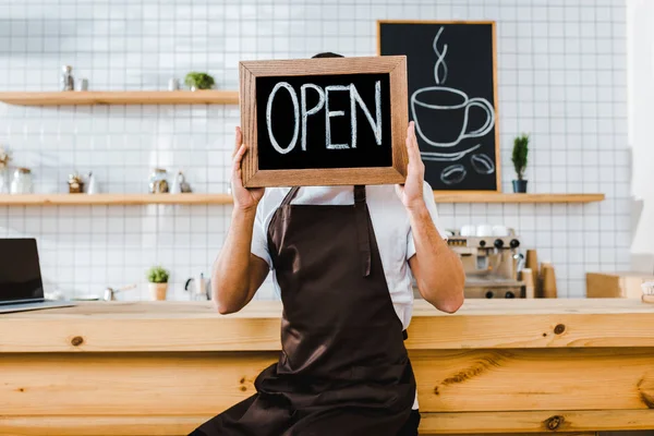 cashier in apron sitting on chair near wooden bar counter and holding chalkboard in front of face with open lettering in coffee house