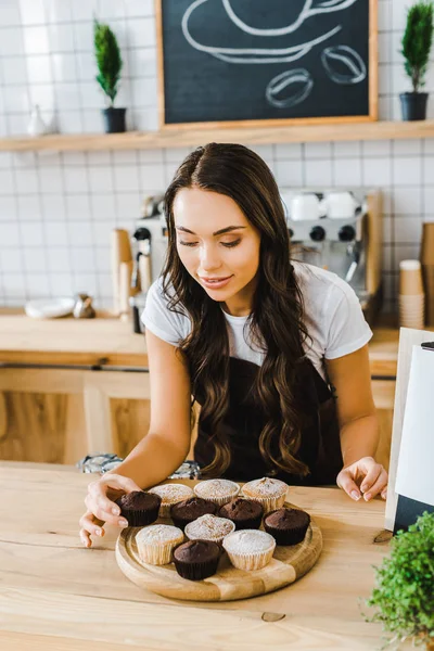 attractive brunette waitress standing behind bar counter and putting cupcakes on wooden tray in coffee house