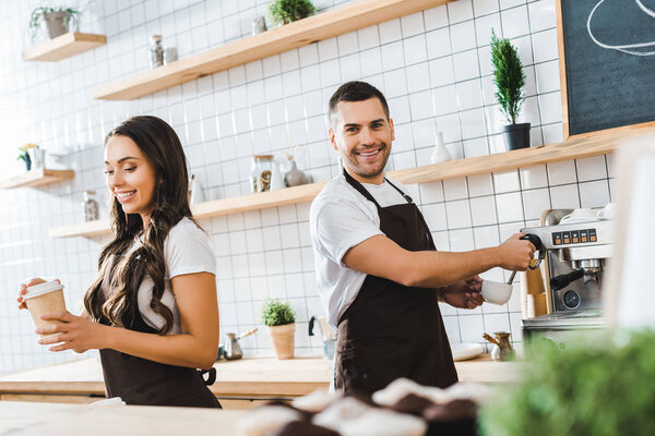 brunette attractive cashier holding paper cup wile barista making coffee in coffee house