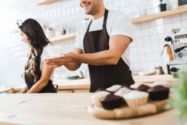 yakışıklı kasiyer Kupası ve fincan tabağı tutan kırpılmış görünümünü cezbetmek çekici esmer barista kahve evde çalışma