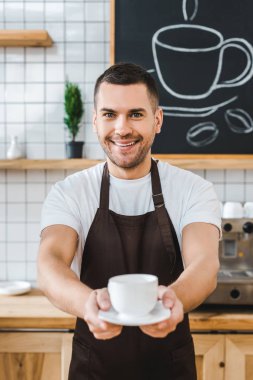 kahverengi önlük Kupası ve fincan tabağı kahve evin içinde tutarak yakışıklı barista seçici odak