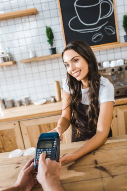 attractive cashier standing near bar counter in brown apron and holding terminal wile man making transaction in coffee house clipart