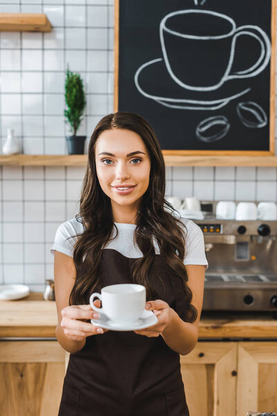 attractive barista in brown apron holding cup and saucer in coffee house