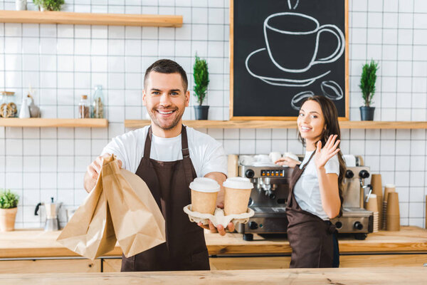 handsome cashier holding paper cups and bags wile attractive brunette barista wavingin in coffee house