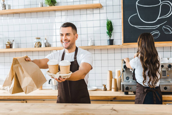 handsome cashier holding paper cups and bags wile attractive brunette barista making coffee in coffee house