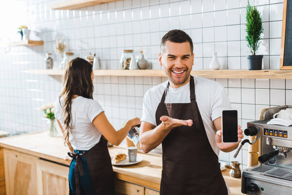 handsome cashier pointing with hand to smartphone with blank screen wile barista working in coffee house