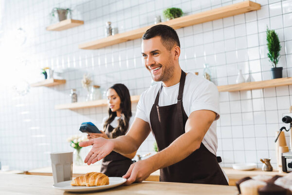 handsome cashier pointing with hand to croissant wile attractive brunette waitress holding terminal in coffee house