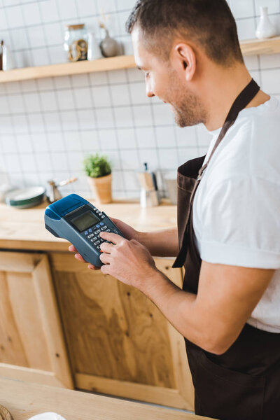 cashier standing in brown apron near bar counter and holding blue terminal in coffee house
