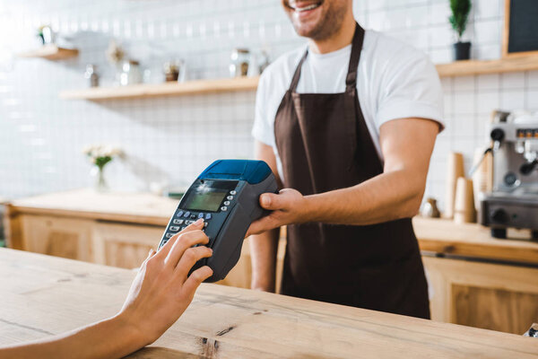 cropped view of cashier standing near bar counter and holding terminal wile woman making transaction in coffee house