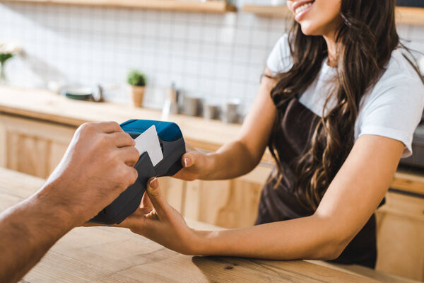 cropped view of cashier standing near bar counter and holding terminal wile man paying with credit card in coffee house