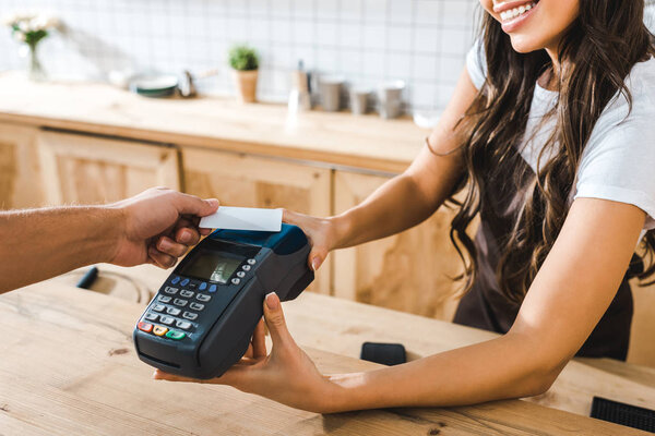 cropped view of cashier standing near bar counter in brown apron and holding terminal wile man paying with credit card in coffee house