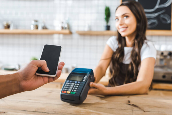 selective focus of male hand holding smartphone wile cashier standing near bar counter and holding terminal in coffee house
