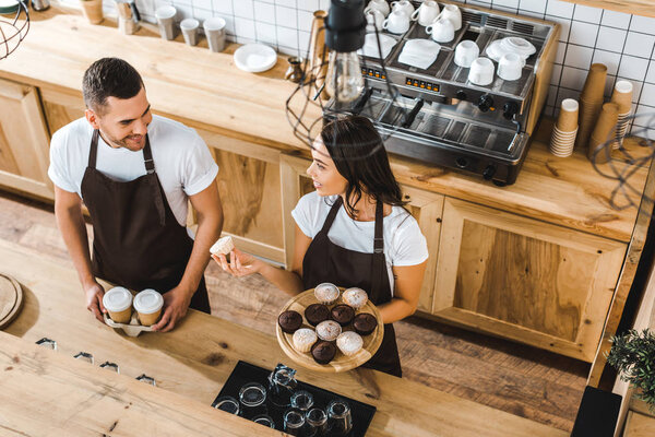 attractive cashier with cupcakes and handsome barista with paper cups standing behind bar counter in coffee house