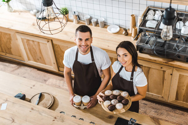 attractive cashier with cupcakes and handsome barista with paper cups standing and smiling in brown aprons behind bar counter in coffee house