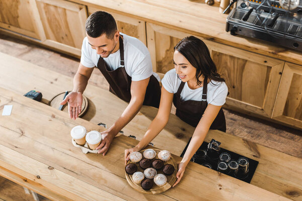 attractive cashier and handsome barista offering cupcakes and coffee in paper cups behind wooden bar counter in coffee house