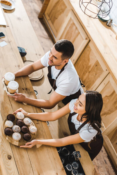 cashiers in brown aprons offering cupcakes and coffee in paper cups behind wooden bar counter in coffee house