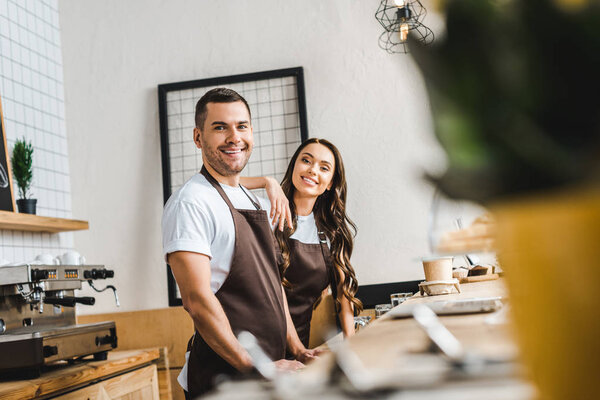 selective focus of cashiers in brown aprons standing and smiling behind wooden bar counter in coffee house